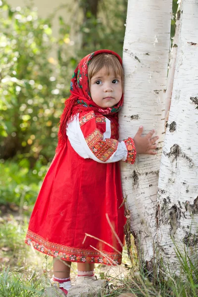 Little girl in Russian traditional dress standing next to a birch