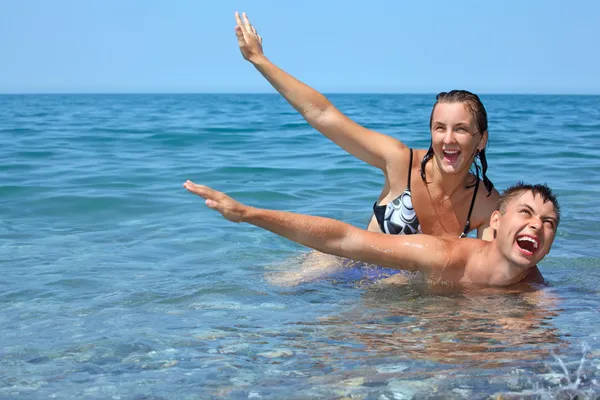 Young woman sitting astride man in sea near coast, lifted hands