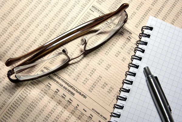 Glasses, pen and notebook laying on newspaper with financial num