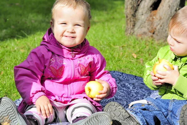 Two small children sit on a green clearing eat apples