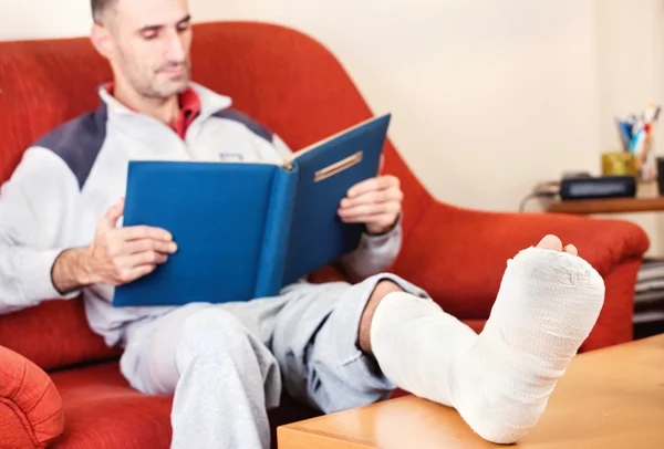 Man with a broken leg on a sofa at home reading book
