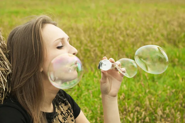 Chica joven sentada en la hierba y soplando burbujas de jabón —  Fotos de Stock