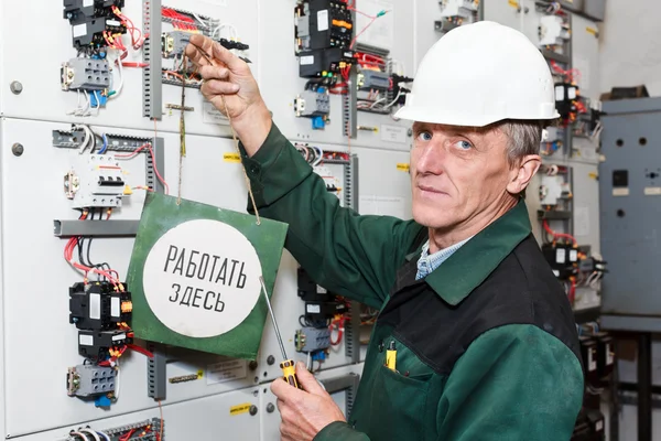 Mature electrician working in white hard hat with caution board — Stock Photo, Image