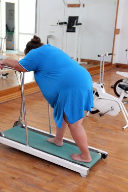 Overweight woman running on trainer treadmill clipart