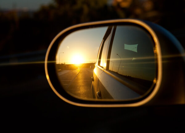 A sunset in the rearview mirror of car as a races down the road