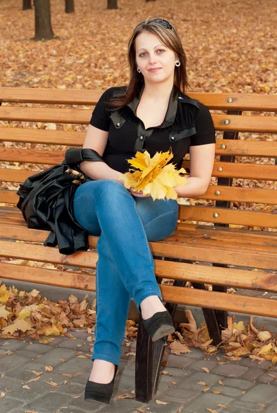 Krásná dívka s podzimní listí, sedí na lavičce v podzimní p — Stock fotografie