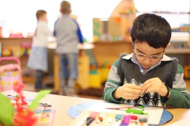 Boy in glasses moulds from plasticine on table in kindergarten clipart