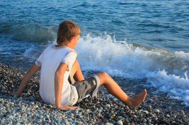 Sitting teenager boy on stone seacoast, wets feet in water, sitt clipart