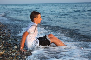 Sitting teenager boy in wet clothes on stone seacoast clipart