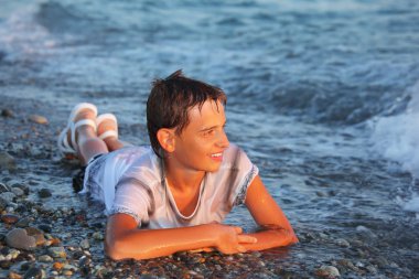 Teenager boy in wet clothes lying on stones on seacoast clipart