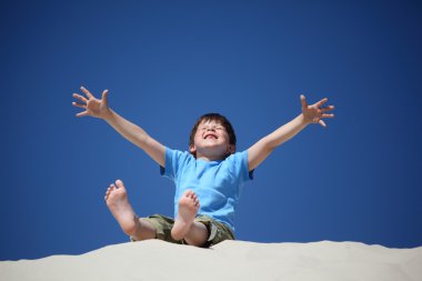 Boy sits on sand with lifted hands with closed eyes clipart