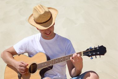 Hasır şapkalı adam kumların üzerinde gitar çalıyor.