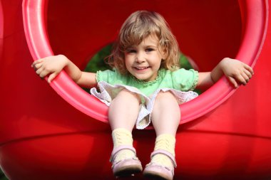 Little girl sits in red plastic hole on playground clipart