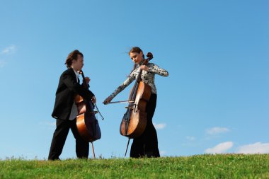 iki violoncellists çim gökyüzü karşı oynamak