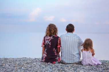 Happy family with little girl sitting on stony beach, sitting by clipart