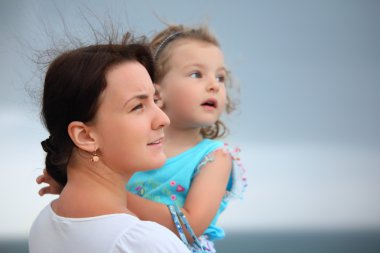 Beautiful woman protects little girl from wind on seacoast clipart