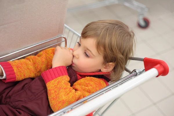 Little girl in shop lies in cart and holds finger in mouth — Stock Photo, Image