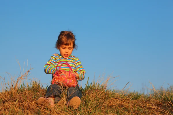 Děvčátko v proužkované tričko s červenou taškou sedí na suché trávy — Stock fotografie