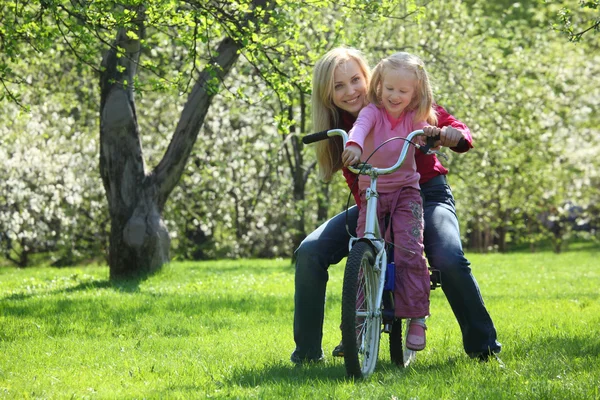 Chica con madre sentarse en bicicleta en floreciente jardín de primavera —  Fotos de Stock