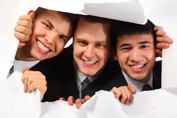Трое молодых улыбающихся мужчин, смотрящих в дыру в бумаге — стоковое фото