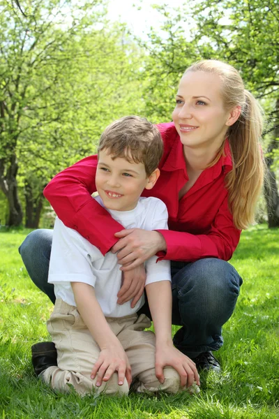 Mother embraces behind son sitting in lap on grass in park in — Stock Photo, Image