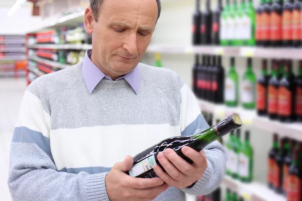 Homem idoso na loja olha em garrafa de vinho em mãos — Fotografia de Stock