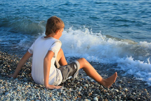 Sitting teenager boy on stone seacoast, wets feet in water, sitt — Stock Photo, Image