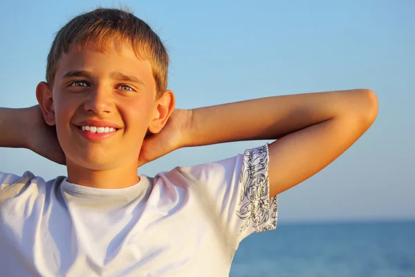 Smiling teenager boy against sea, holding hands behind head — Stock Photo, Image
