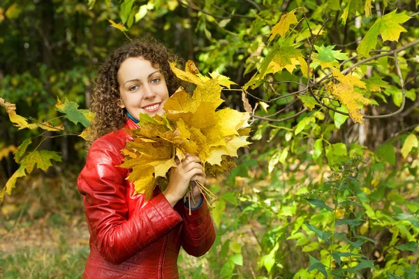 Mujer joven con hojas de arce en las manos cerca del árbol en madera en autu —  Fotos de Stock