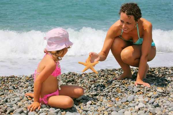 Ung kvinna ger sjöstjärnor till liten flicka på stenig strand — Stockfoto