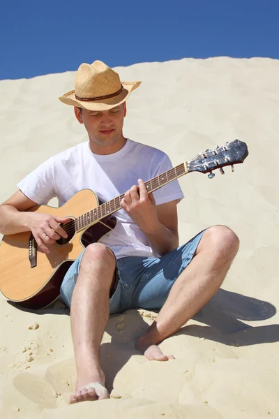 Ο τύπος με το ψάθινο καπέλο παίζει κιθάρα καθισμένος στην άμμο — Φωτογραφία Αρχείου