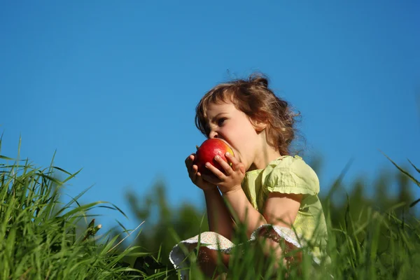 Dziewczyna zjada czerwone jabłko w trawie przeciw błękitne niebo — Zdjęcie stockowe