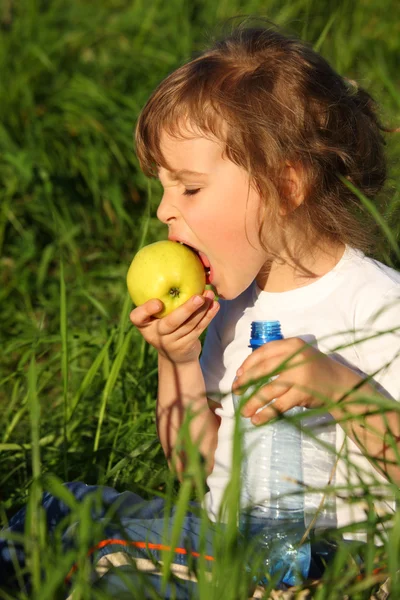 Menina com garrafa de plástico come maçã verde na grama — Fotografia de Stock