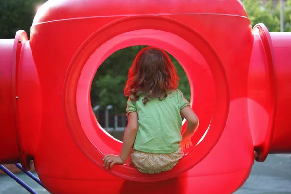 Niña se sienta en caja de plástico rojo en el patio de recreo, vista desde bac —  Fotos de Stock