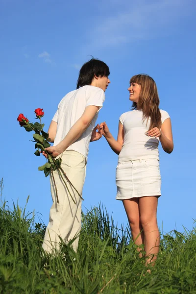 Young pair stands on a grass, guy holds behind back bouquet of — Stock Photo, Image