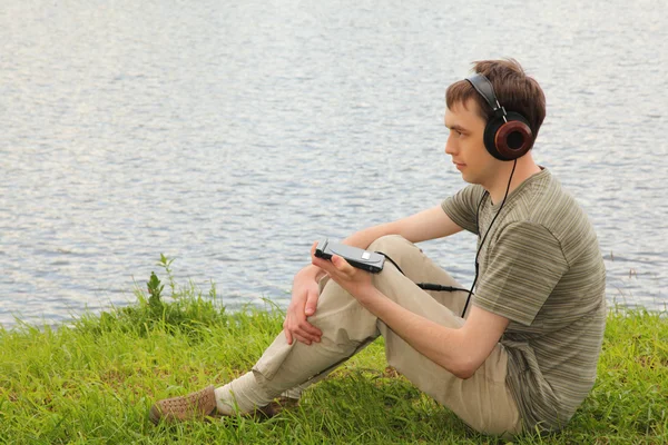Молодой человек увязывает музыку в наушниках сидит на траве на берегу — стоковое фото