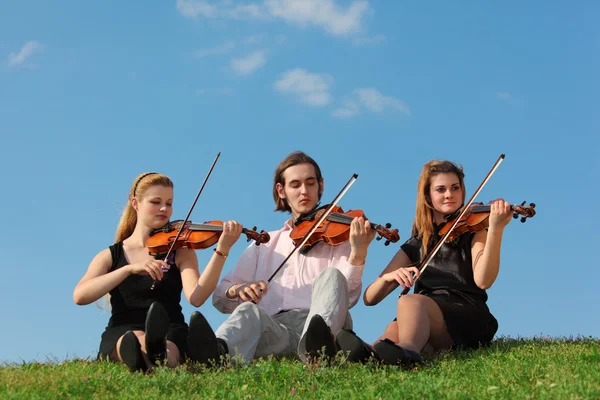 Three violinists sit and play on grass against sky — Stock Photo, Image