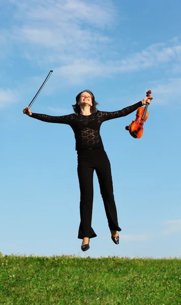Meisje met viool springt op gras tegen hemel — Stockfoto