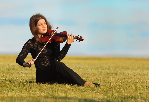 Meisje zit op gras en viool speelt tegen hemel — Stockfoto