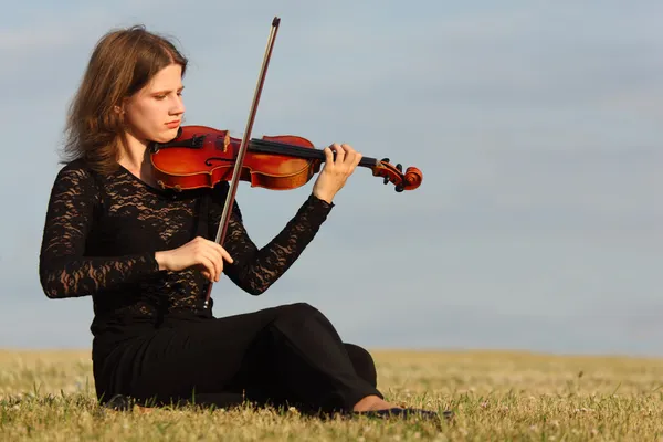 少女は草の上に座っているし、空を背景にバイオリンを演奏 — ストック写真