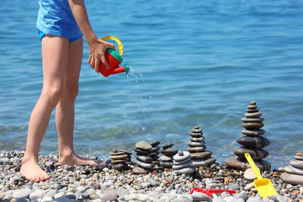 Child waters from watering can stone stacks on beach — Stock Photo, Image