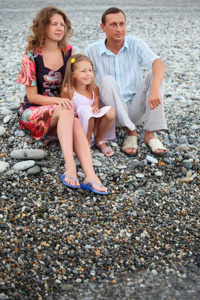 Happy family with little girl sitting on stony beach — Stock Photo, Image