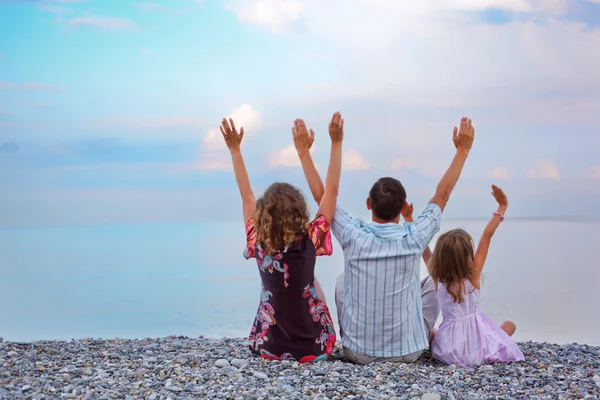 Happy family with little girl sitting on stony beach lifted hand — Stock Photo, Image