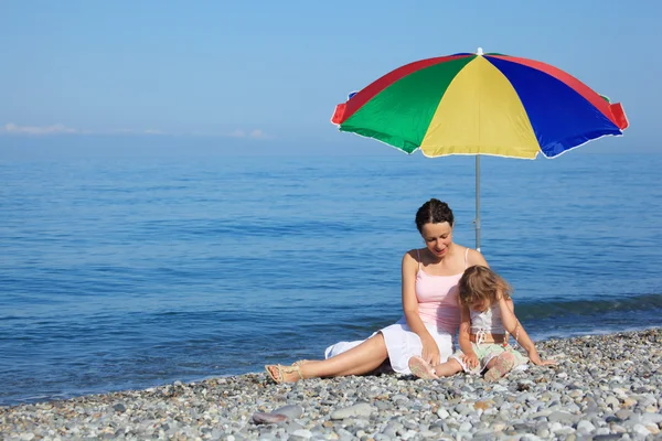 Мать с ребенком под разноцветным зонтиком на галечном пляже — стоковое фото