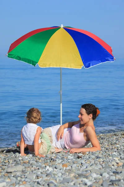 与在卵石海滩上多彩色伞下的孩子的母亲 — 图库照片