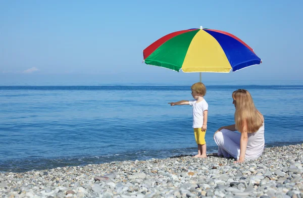 Μητέρα με το παιδί κάτω από ένα πολυ χρωματισμένη ομπρέλα στην παραλία με βότσαλα — Φωτογραφία Αρχείου