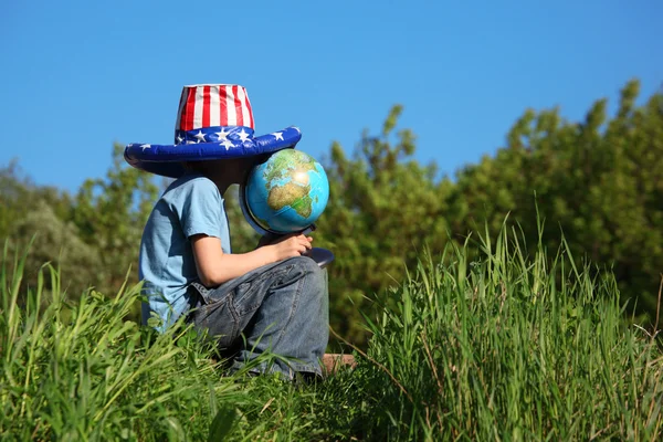 Мальчик в большой шляпе с американским флагом сидит на траве и держит в руках глобус — стоковое фото