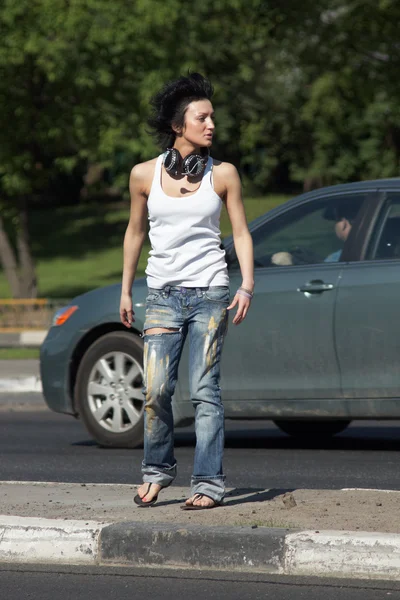 Girl with ear-phones stands on road among cars — Stock Photo, Image