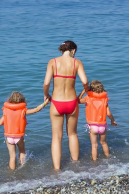 Two little girls in lifejackets with young woman coming into sea clipart