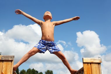 Boy in shorts standing on boards against clouds, plants hands an clipart